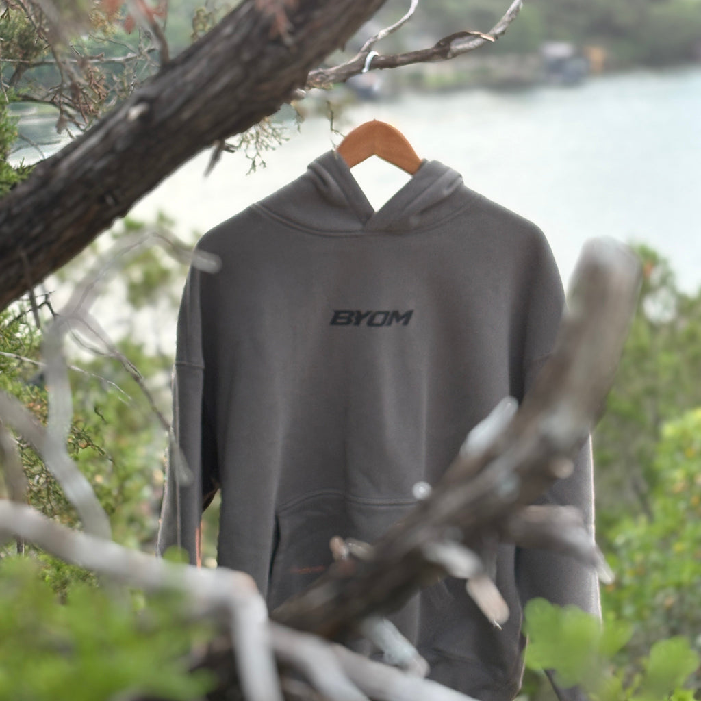 Gray hoodie with 'BYOM' logo hanging on a tree branch by a body of water.